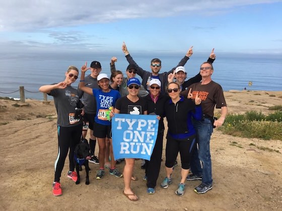 Group of about twelve runners with a dog posing on a coastal hilltop overlooking the ocean, arms raised in celebration