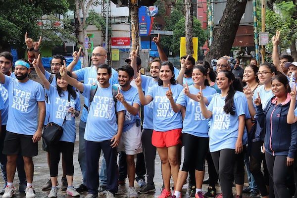 Enthusiastic crowd of runners in light blue shirts cheering and clapping while walking together along a tree-lined city street