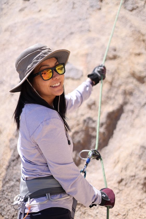 A smiling woman in reflective sunglasses and a wide-brimmed hat rock climbing on a sunlit rock face with rope gear