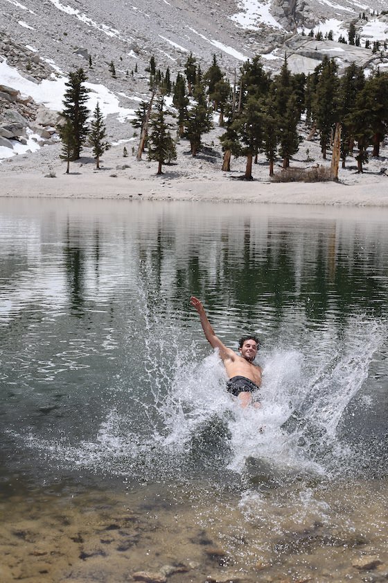 A person splashing joyfully into a cold alpine lake surrounded by snow-dusted mountains and evergreen trees
