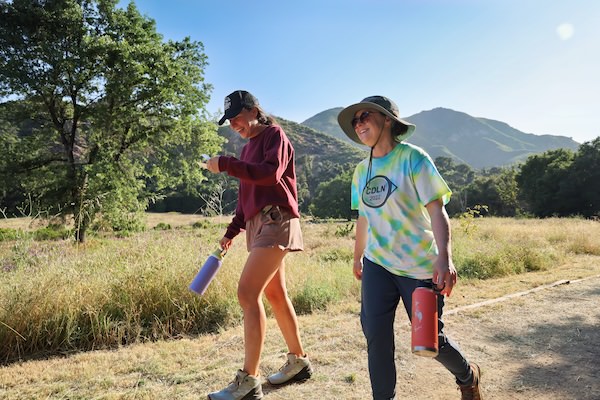 Two women walking and smiling along a sunny grassy trail with green mountains rising in the background